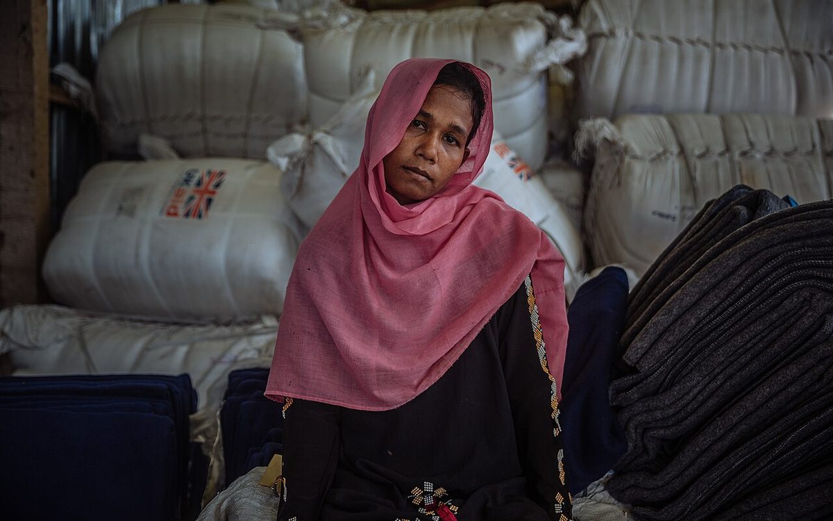 A Rohingya woman in a pink headscarf seated among sacks and bales of relief supplies